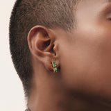 Primer plano editorial de unos aretes tipo huggie elaborados en bronce con baño en oro, engastados con esmeraldas colombianas en bruto seleccionadas por su forma orgánica y su intensidad de color. Su tamaño compacto y su diseño delicado los convierten en una pieza esencial, perfecta para el uso diario con un toque de sofisticación natural. La imagen resalta un trabajo artesanal cuidadoso y la pureza del verde característico de Fenomena.