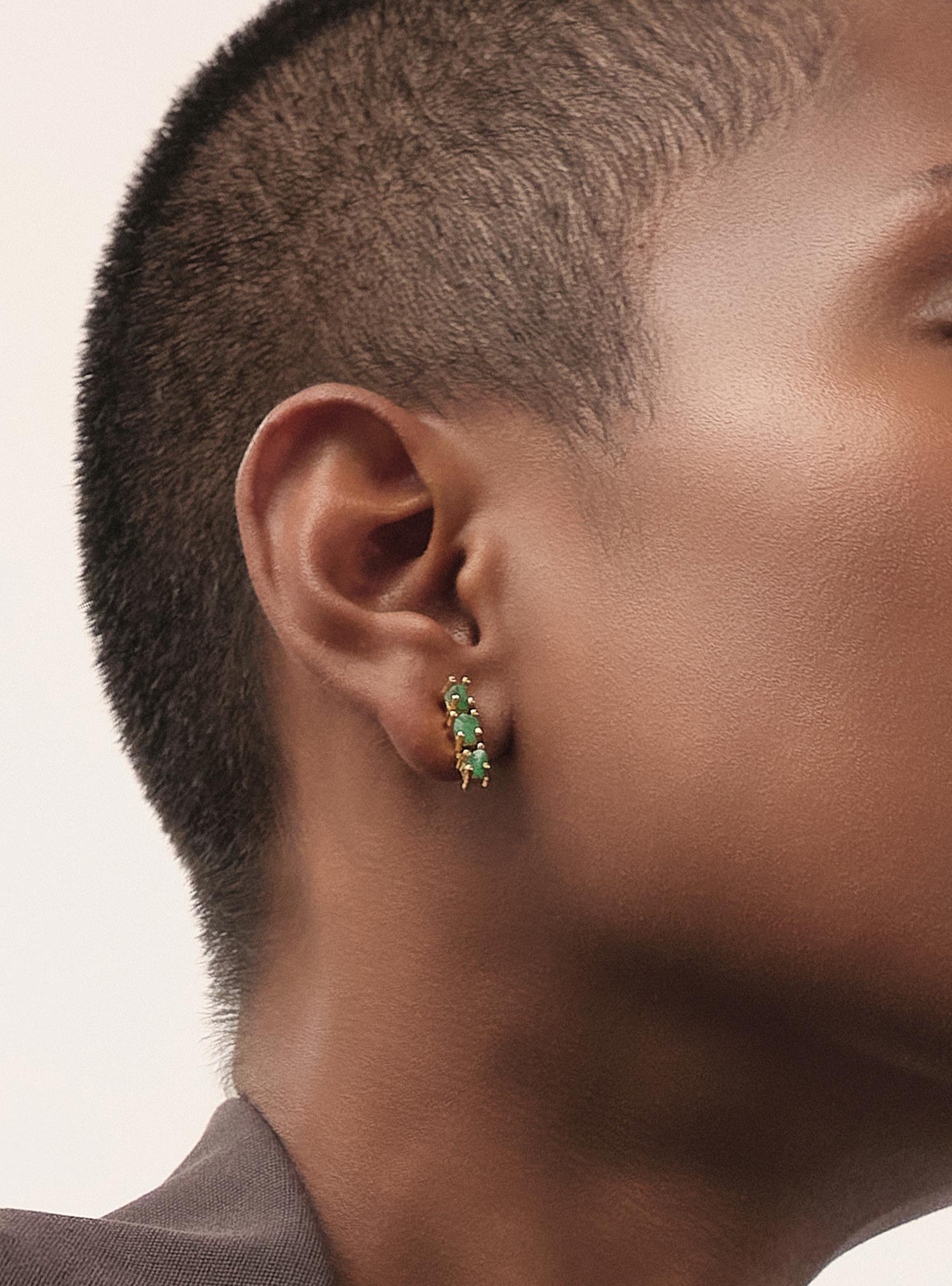 Primer plano editorial de unos aretes tipo huggie elaborados en bronce con baño en oro, engastados con esmeraldas colombianas en bruto seleccionadas por su forma orgánica y su intensidad de color. Su tamaño compacto y su diseño delicado los convierten en una pieza esencial, perfecta para el uso diario con un toque de sofisticación natural. La imagen resalta un trabajo artesanal cuidadoso y la pureza del verde característico de Fenomena.