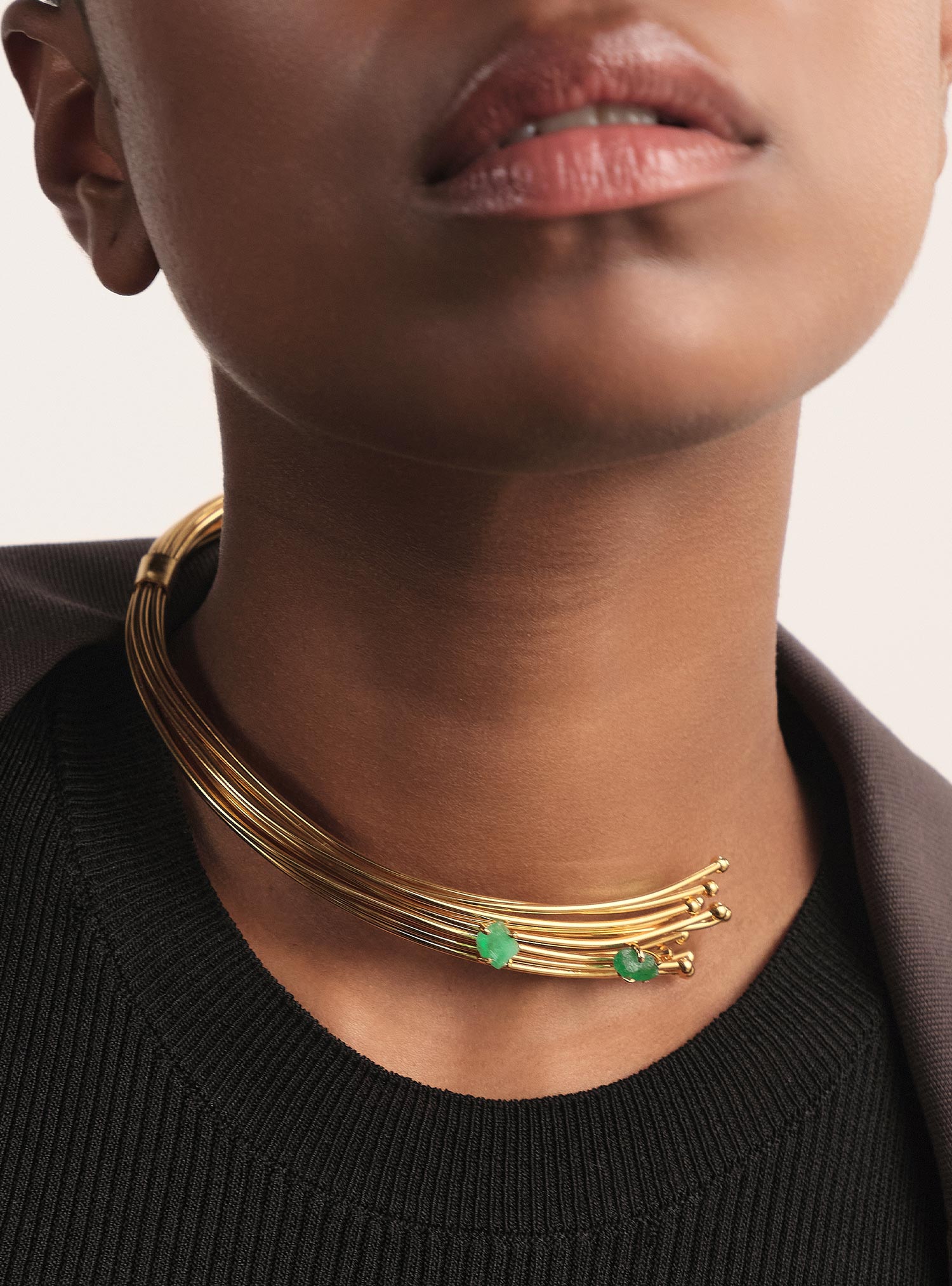 Close-up editorial de un collar de alta joyería compuesto por múltiples hilos de bronce bañados en oro, cuidadosamente ensamblados y terminados a mano. Dos esmeraldas colombianas en bruto, seleccionadas por su forma orgánica y su rareza, se integran de manera sutil y precisa en la pieza, aportando un acento verde puro que eleva el diseño. La imagen destaca el refinamiento del acabado, la delicadeza del volumen y la esencia minimalista de un collar exquisitamente trabajado por Fenomena.